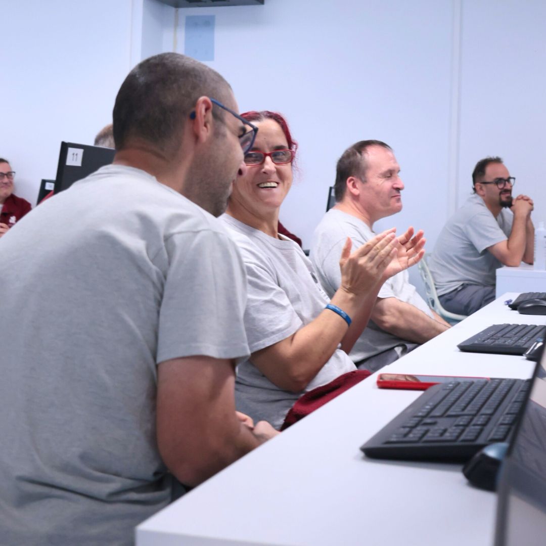 Grupo de personas en una charla frente varios escritorios; una de ellas está de espaldas a la cámara, otra aplaudiendo mientras la mira, y el resto de las personas miran al frente