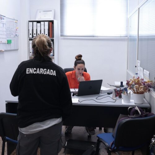 Persona trabajadora de Asprona concentrada en el ordenador en su escritorio, con una persona encargada de espaldas frente a ella en una oficina.