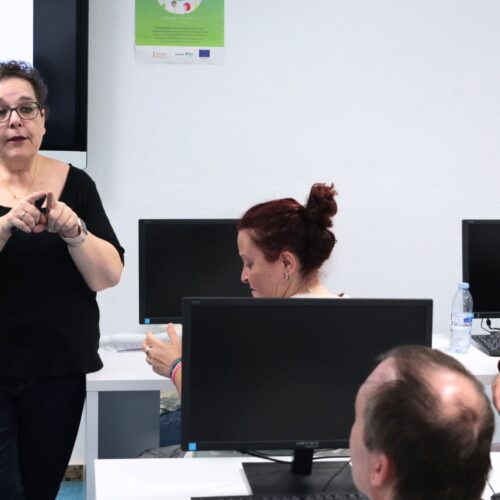Persona de pie dando una presentación frente a otras personas sentadas en escritorios con ordenadores, prestando atención a la presentación