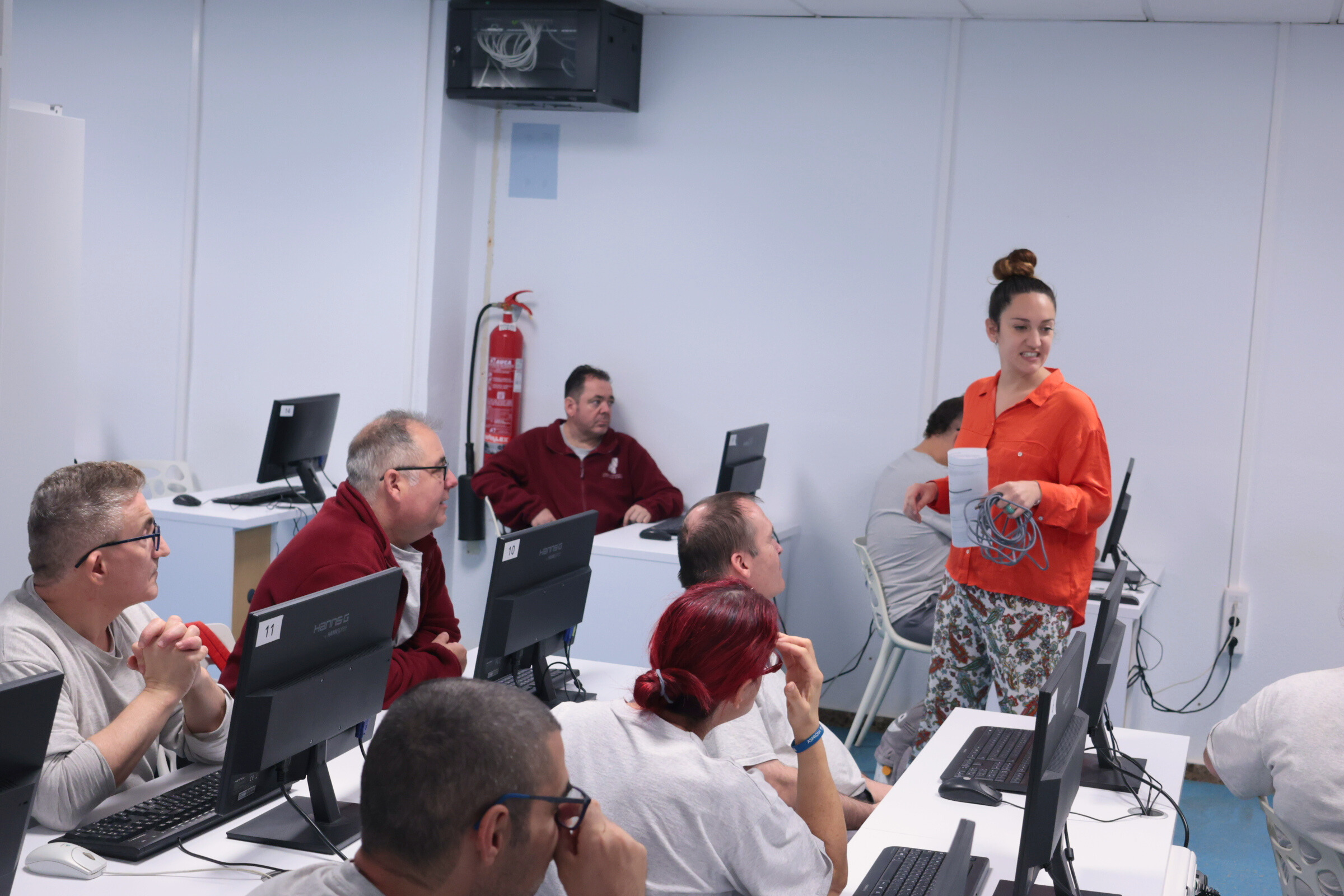Persona de pie, sonriente, dando una presentación, mientras otras personas sentadas en escritorios con ordenadores prestan atención